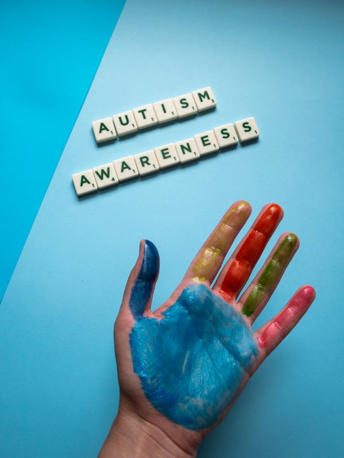 A top view of a colorful painted hand next to autism awareness scrabble tiles on a blue background.