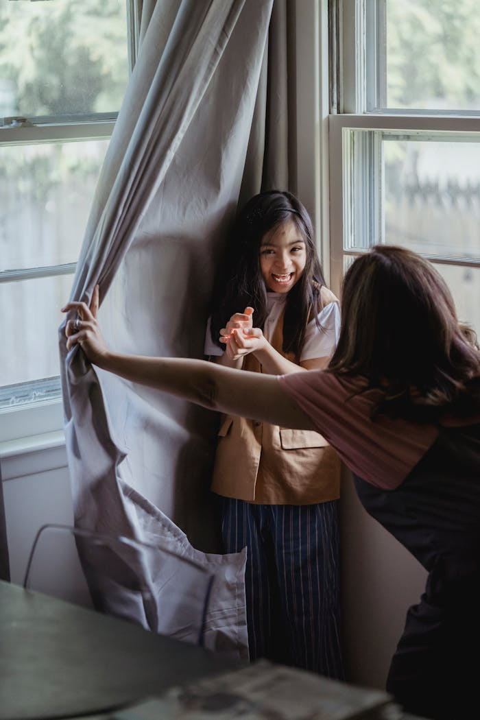A mother and daughter enjoy a playful moment playing hide and seek by the window indoors.